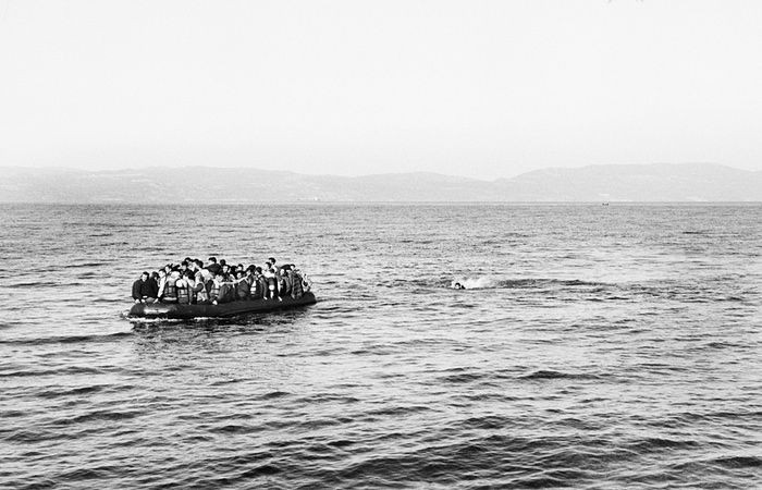 Một con thuyền chật ních người di cư đang hướng vào bờ. Cách đó không xa, một người Syria đã bị rơi khỏi thuyền và đang chờ cứu hộ.