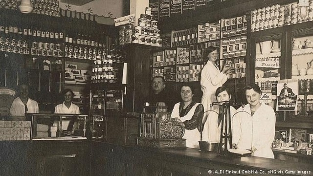 Theo Albrecht Jr.  Cửa hàng bán lẻ trong bức ảnh được thành lập năm 1913 gần Essen, Đức. Tồn tại xuyên suốt thời kỳ siêu lạm phát của nền Cộng hòa Weimar và sự sụp đổ của Đức Quốc xã, cửa hàng này cuối cùng đã phát triển trở thành chuỗi siêu thị chiết khấu lớn nhất của Đức, Aldi. Theo Albrecht Jr. là con trai của Karl và Anna Albrecht, từng sở hữu một cửa hàng thực phẩm ở góc đường trước khi các cậu con trai mở rộng nó thành đế chế Aldi như hiện nay.
