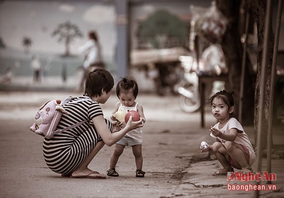 Ba mẹ con trên đường đi học về. Đứa em út đang tuổi tập đi, nhõng nhẽo đòi mẹ chơi đồ chơi, người mẹ chiều con ngồi xuống bên vệ đường 