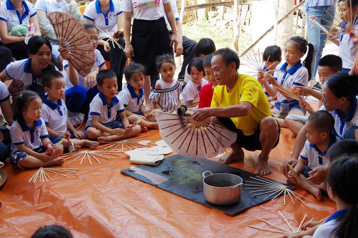 Các bé chăm chú lắng nghe và quan sát nghệ nhân làng Nam giới thiệu, thực hành các công đoạn làm quạt giấy. Ảnh: Phương Linh