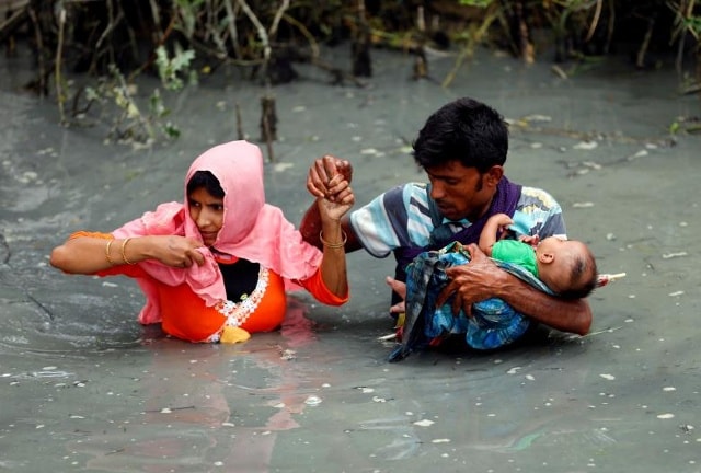 Những người tị nạn Rohingya mang con của họ khi họ đi bộ qua nước sau khi băng qua biên giới bằng thuyền qua sông Naf ở Teknaf, Bangladesh.