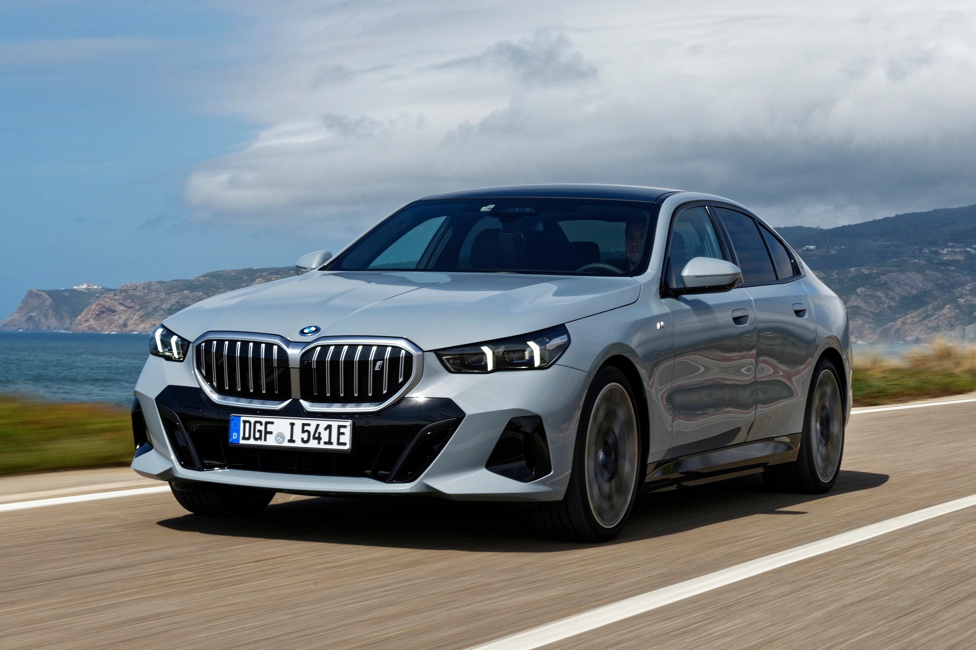 Chiếc BMW i5 màu xanh di chuyển trên đường ven biển, thể hiện thiết kế hiện đại của hãng xe Đức.